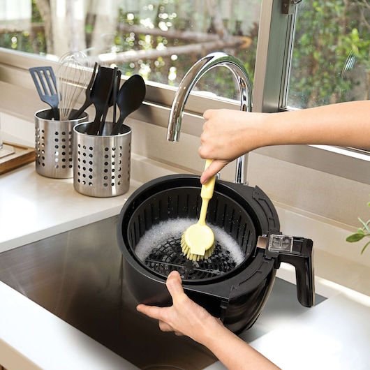 Photo of hands cleaning BLACKDECKER air fryer AF400 basket with a yellow brush by a kitchen sink.