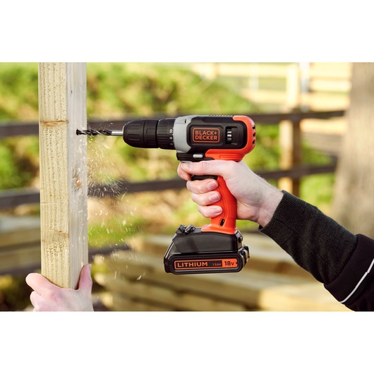 BLACK+DECKER BCD001C2K cordless drill used to drill into wood outdoors, orange and black design, visible 18V lithium battery.