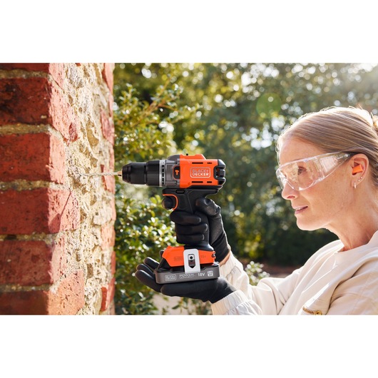Person using BLACK+DECKER cordless drill to drill into a brick and stone wall outdoors, wearing safety glasses.