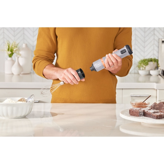 BLACKDECKER BCKM101WHFF hand mixer held in kitchen scene, whisk attachment, baking ingredients visible.