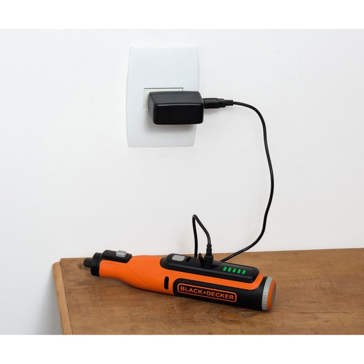 BLACK+DECKER BCRT8I cordless rotary tool charging on a wooden surface with adapter plugged into a wall socket.