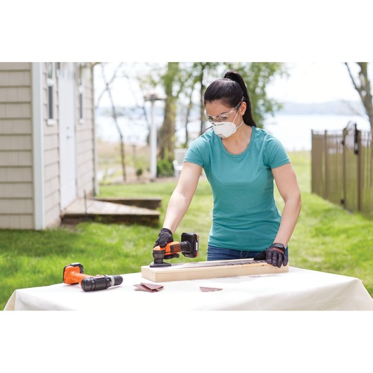 Photo of person using BLACKDECKER BDCK502C1 cordless sander on wood outdoors, drill and sandpaper on table.
