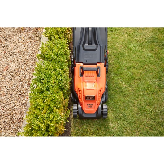 Photo of BLACK+DECKER BEMW471BH electric lawn mower cutting grass, top view, orange and black design beside hedge.