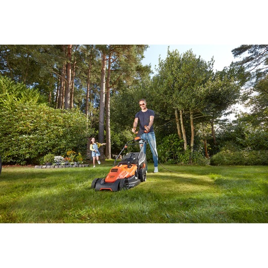 BLACKDECKER BEMW481ES lawn mower in use on grass in a garden with trees and a child in the background.