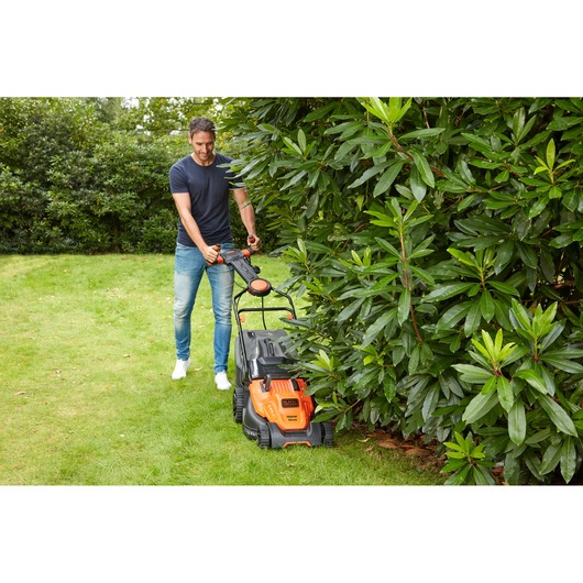 BLACK+DECKER BEMW481ES lawn mower in use on grass near bushes, orange and black design, outdoor setting.