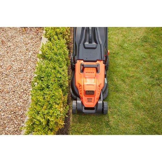 Top view of BLACK+DECKER BEMW481ES lawn mower with orange and black design mowing grass near hedge.