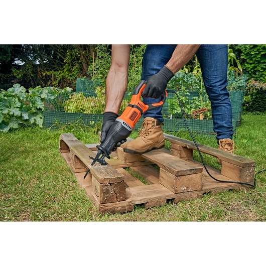 A person wearing gloves and work boots is using a BLACK+DECKER reciprocating saw to cut through a wooden pallet outdoors on grass, with a garden in the background.