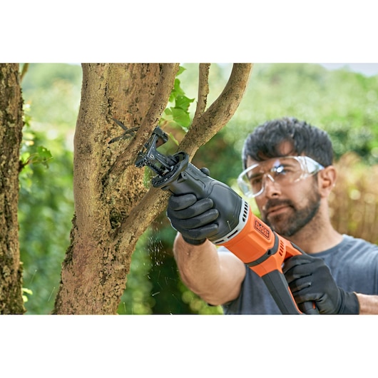 A person wearing gloves uses a BLACK+DECKER reciprocating saw to cut a tree branch outdoors. The saw is orange and black, and sawdust is visible as the branch is being cut.