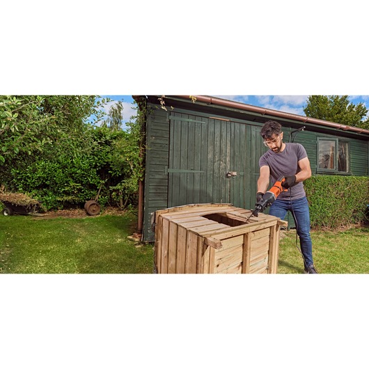 A person using a BLACK+DECKER electric reciprocating saw (model BES301K) to cut a wooden crate outdoors, wearing gloves and standing near a green garden shed.