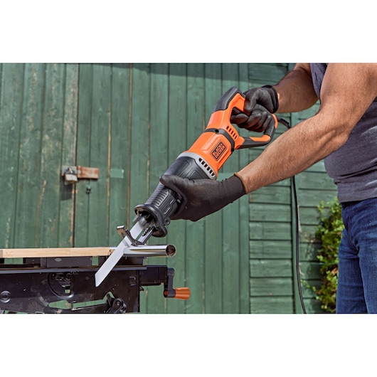 A person wearing gloves is using a BLACK+DECKER reciprocating saw to cut a metal pipe, with the saw positioned on a workbench outdoors.