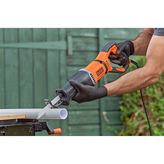 A person using a BLACK+DECKER reciprocating saw to cut a white plastic pipe outdoors. The saw is orange and black and held with gloved hands.