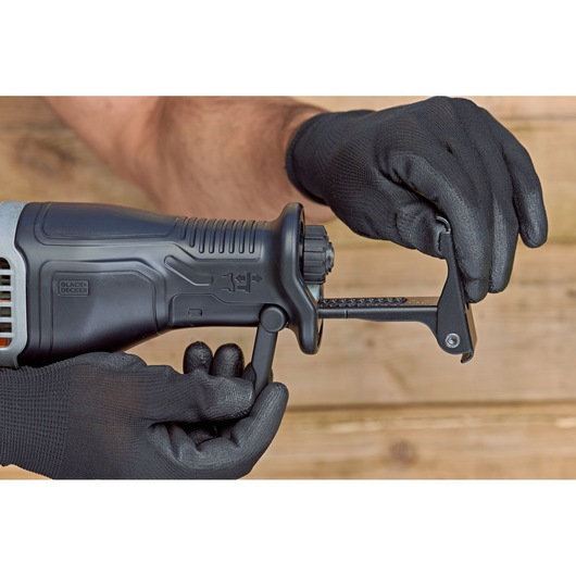 Close-up of gloved hands adjusting the blade on a BLACK+DECKER reciprocating saw, shown against a wooden background.