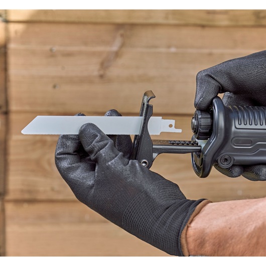 Close-up of gloved hands installing a reciprocating saw blade into a BLACK+DECKER power tool, with a wooden background.