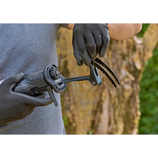 Close-up of a person wearing black gloves and assembling or adjusting a BLACK+DECKER electric tool with a claw attachment outdoors, in front of a tree trunk.