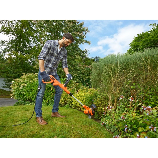 BLACKDECKER BESTA530CM grass trimmer being used in a garden, orange and black design, outdoor setting.