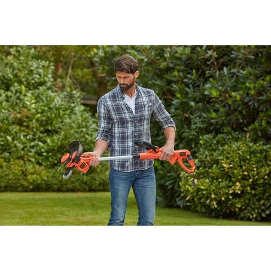 Photo of person holding BLACKDECKER BESTA530CM orange grass trimmer with cord in a garden setting.