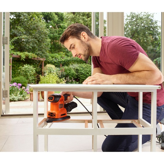BLACKDECKER BEW200 sander in use sanding a white wooden table indoors near open doors.