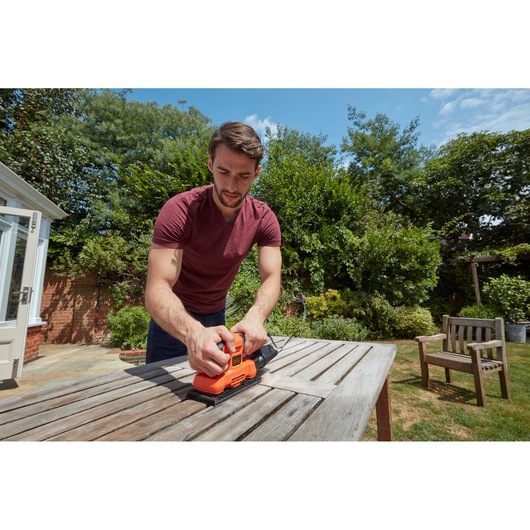Person using BLACKDECKER BEW220-GB sander on outdoor wooden table in garden setting.