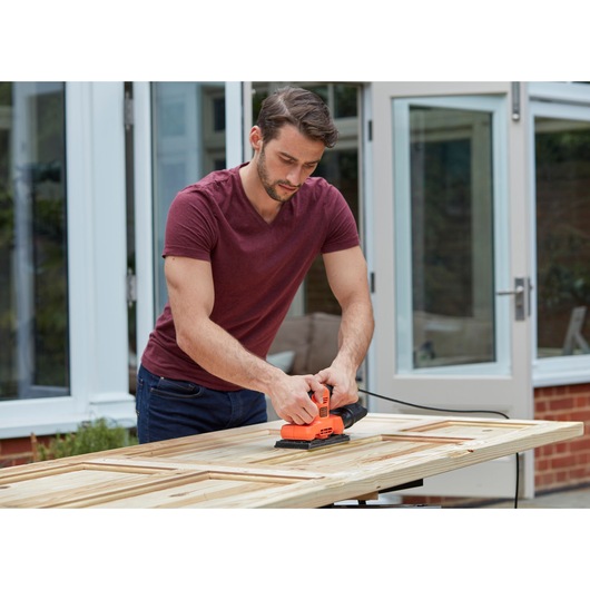 Person using BLACK+DECKER BEW220-GB orange electric sander on wooden door outdoors.