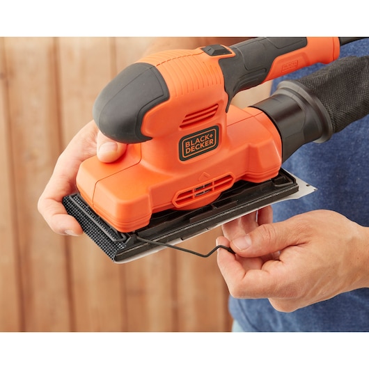 BLACK+DECKER BEW220-GB orange detail sander shown in hand, close-up of sandpaper attachment mechanism.