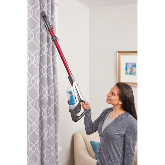 BLACK+DECKER BHFE620J cordless stick vacuum with red tube cleaning patterned grey curtains in living room.