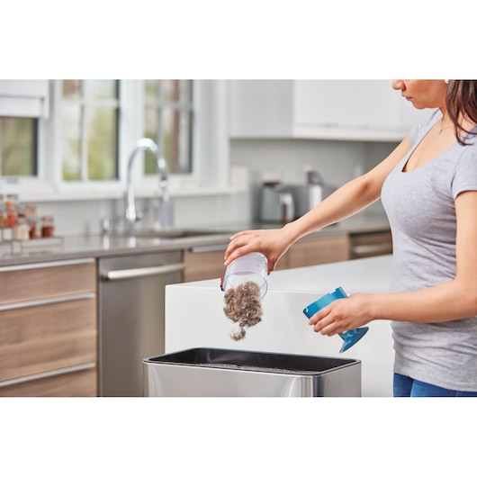 Person emptying BLACKDECKER BHFE620J vacuum dust container into trash bin in modern kitchen.