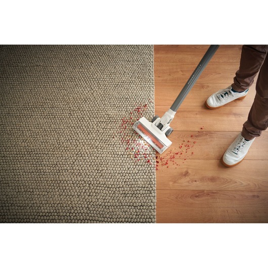 A person uses a BLACK+DECKER cordless vacuum cleaner to clean red debris from the edge of a rug onto a wooden floor.