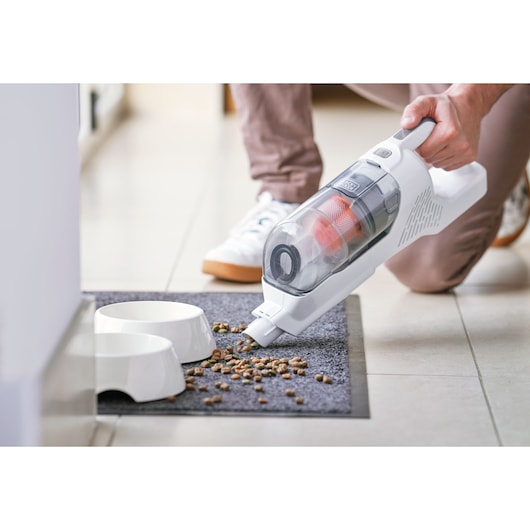 A person is using a cordless handheld vacuum cleaner to pick up scattered pet food from a mat next to two white pet bowls.
