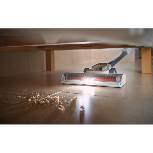 A close-up view of a BLACK+DECKER cordless vacuum cleaner with a lit brush head, cleaning crumbs from a tiled floor beneath furniture.