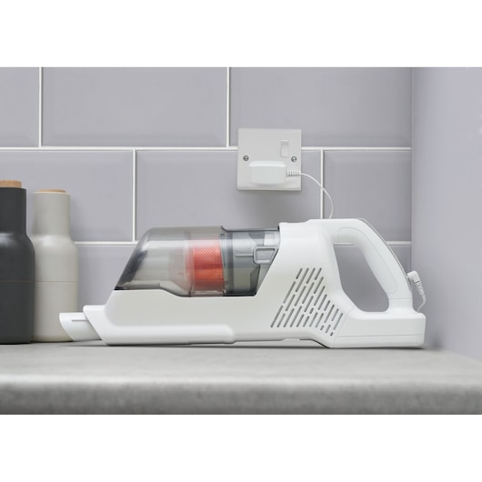 A cordless handheld vacuum cleaner in white, placed on a countertop next to kitchen containers, with a charging cable plugged into a wall socket.