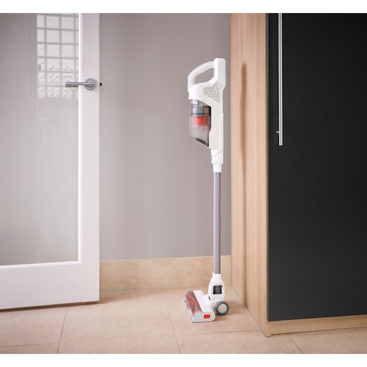 A white cordless stick vacuum cleaner with a transparent dust container is standing upright against a wall in a modern kitchen setting.