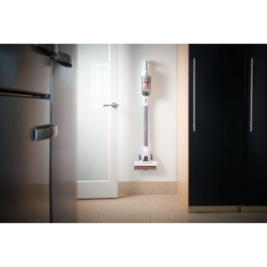 A BLACK+DECKER stick vacuum cleaner is mounted on a white wall in a modern kitchen, positioned between a refrigerator, a cabinet, and a glass door.