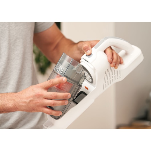 A person is removing the dust container from a BLACK+DECKER cordless vacuum cleaner.