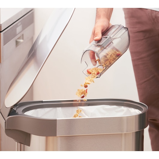 A person empties crumbs and food debris from a transparent vacuum cleaner dust container into a stainless steel trash bin in a kitchen.