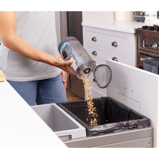 BLACKDECKER BHFEV36B2D vacuum dust canister being emptied into kitchen trash bin, indoor setting.