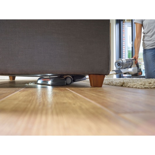 BLACKDECKER BHFEV36B2D cordless vacuum cleaning under sofa on wooden floor in living room.