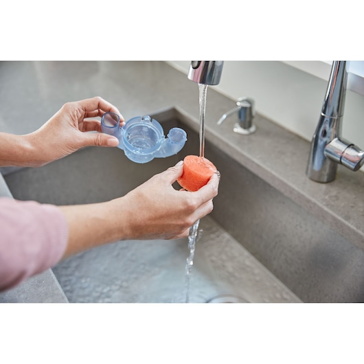 BLACKDECKER BHSB315J hand washing orange sponge under faucet, holding plastic part, kitchen sink visible.