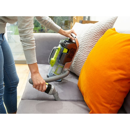 BLACKDECKER CUA525BHA vacuum cleaner used on a grey sofa near orange pillow, side angle.