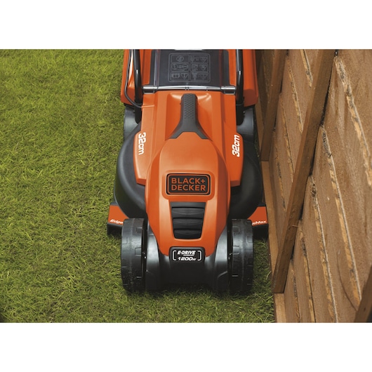 Top view of a BLACK+DECKER electric lawn mower, model EMAX32S, with a 32 cm cutting width, shown mowing grass closely along a wooden fence.
