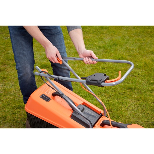A person is adjusting the handle of an orange and black BLACK+DECKER electric lawn mower on grass.