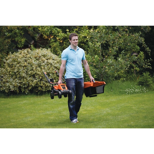 A person in a light blue shirt and jeans carries a BLACK+DECKER EMAX32S electric lawn mower and a black and orange grass collection box across a grassy lawn, with trees and bushes in the background.