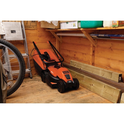 BLACKDECKER EMAX32S lawn mower in orange parked in a wooden shed beside planks and a ladder.