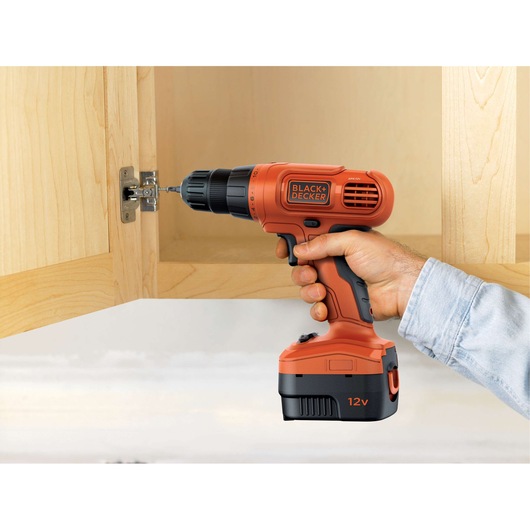 Photo of BLACK+DECKER EPC12CAB cordless drill in use on a cabinet hinge, orange and black design, 12V battery.