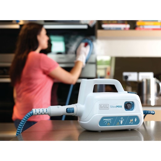 BLACK+DECKER FSH10SMP steam cleaner unit in foreground, woman cleaning oven in kitchen background.
