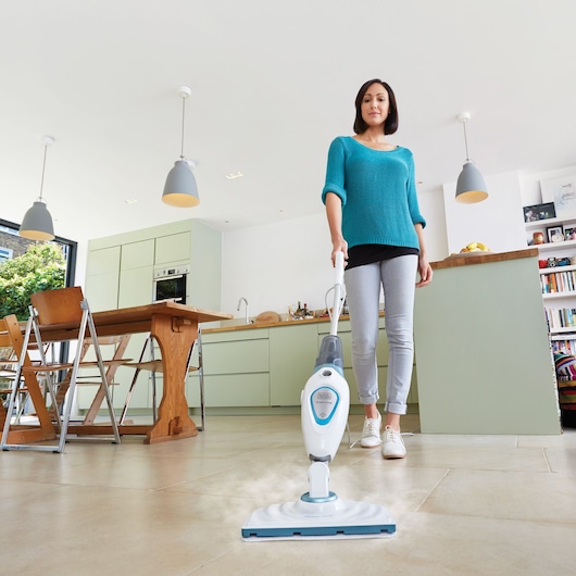 A person with a blurred face uses a BLACK+DECKER steam mop FSM1605 to clean the tiled floor in a modern kitchen. The mop emits steam while being pushed across the floor.