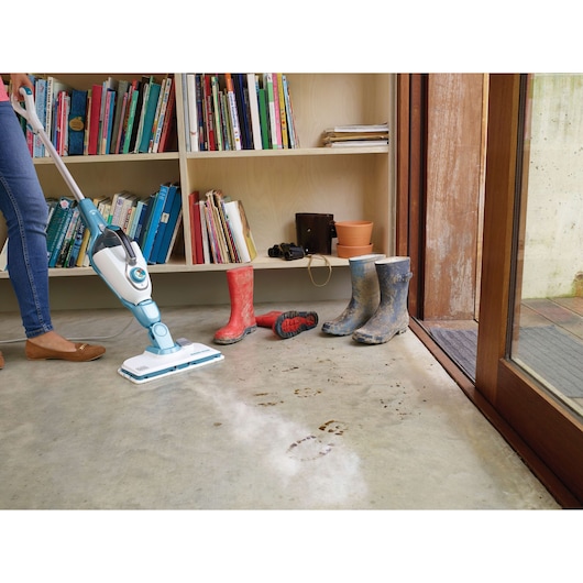 A person uses a BLACK+DECKER steam mop to clean muddy footprints off a concrete floor near a bookshelf and pairs of dirty boots.
