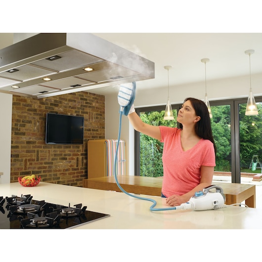 A person using a BLACK+DECKER steam cleaner to clean a kitchen extractor hood. The steam cleaner is white with blue accents and is emitting steam during the cleaning process.
