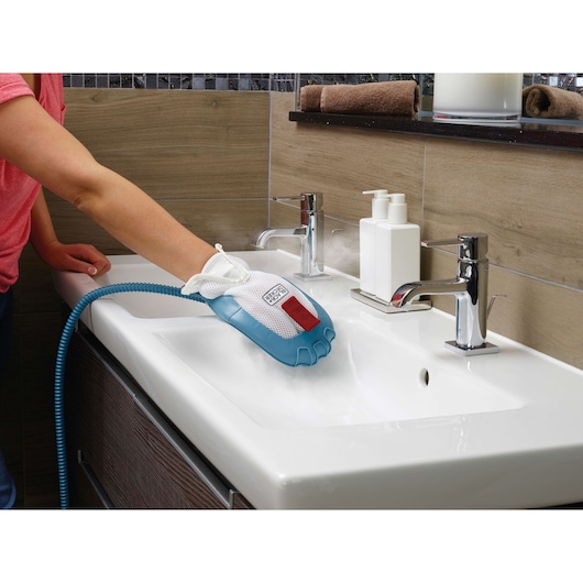A person is using a BLACK+DECKER steam cleaning glove to sanitize a bathroom sink. The glove is connected to a hose and emits steam, cleaning the surface around the faucet.