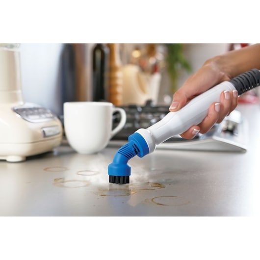 A hand using a BLACK+DECKER steam cleaner with a blue brush attachment to clean coffee stains from a kitchen countertop. A white mug and kitchen appliances are visible in the background.