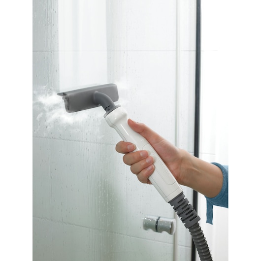 A hand holds a BLACK+DECKER steam cleaning device with a gray nozzle attachment, directing steam onto a tiled glass shower door for cleaning.